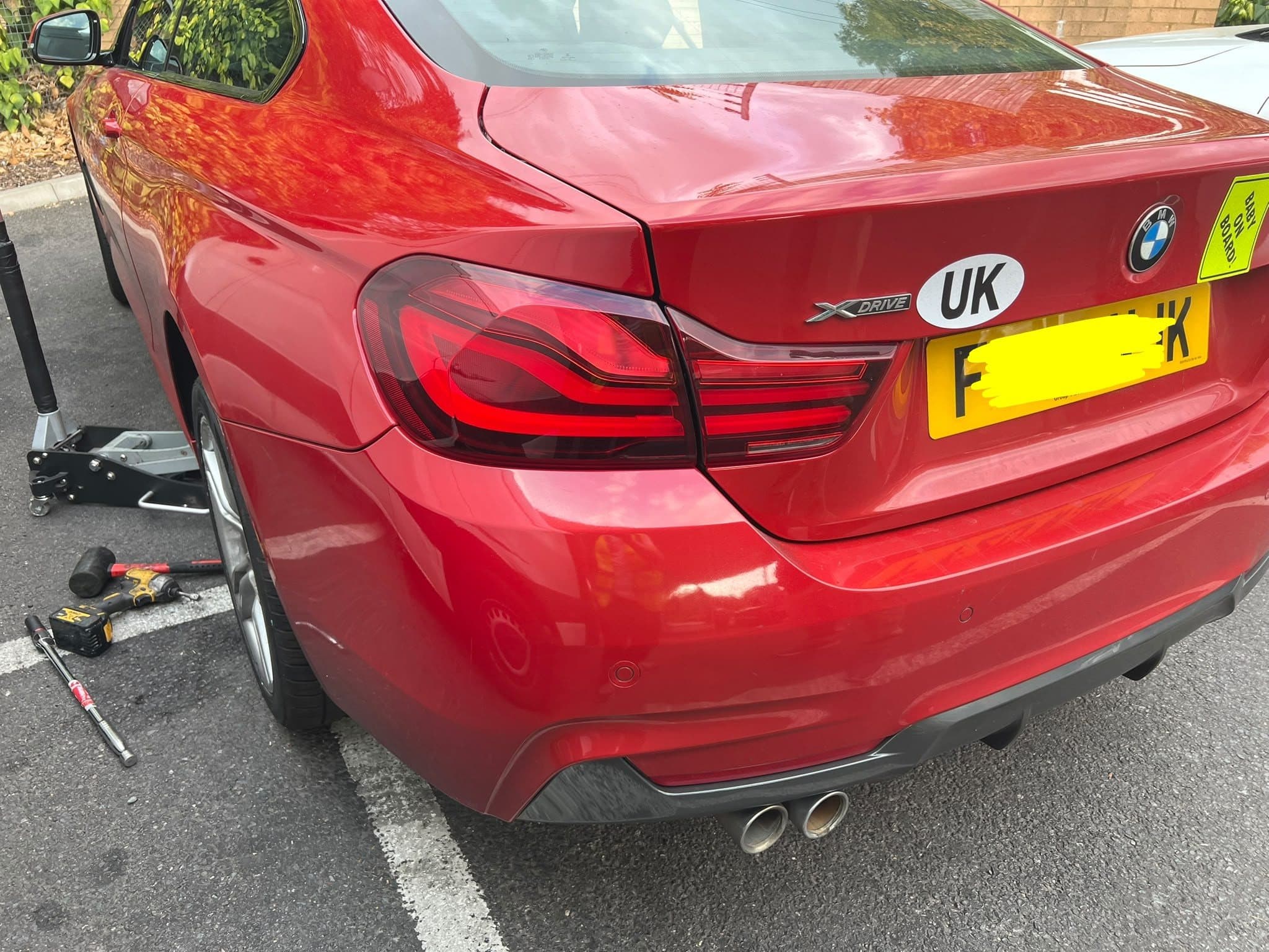 A red BMW coupé with its rear wheel being changed, a trolley jack and an impact driver on the bay beside it