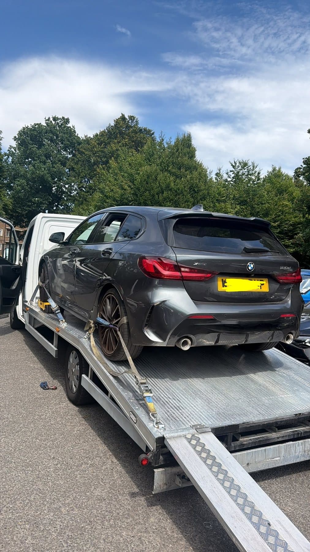 A grey BMW hatchback strapped at the wheels on the bed of a recovery truck, loading ramps still down